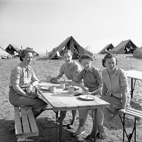 Four women sit at a wooden table in a desert. Several large tents are in the background.