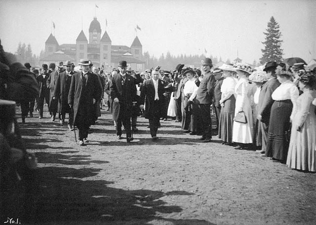 Dignitaries walking past a large crowd. An ornate building is in the background.