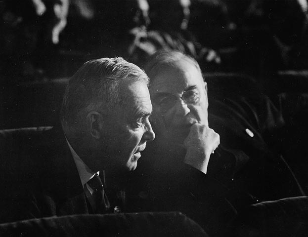 Two middle-aged men in suits lean together and talk while sitting in plush chairs.