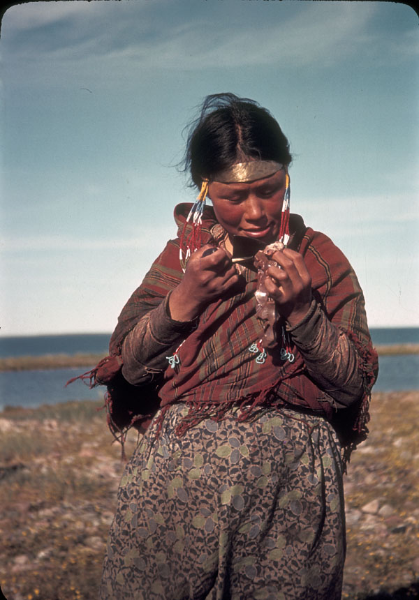 A young Inuit woman looks at a copper necklace in her grip. Behind her is a large body of water.