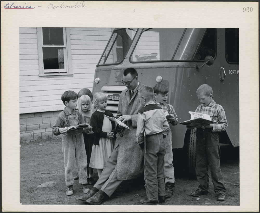 Kids gather around a man holding a book, leaning against the hood of a van. Some kids hold books.