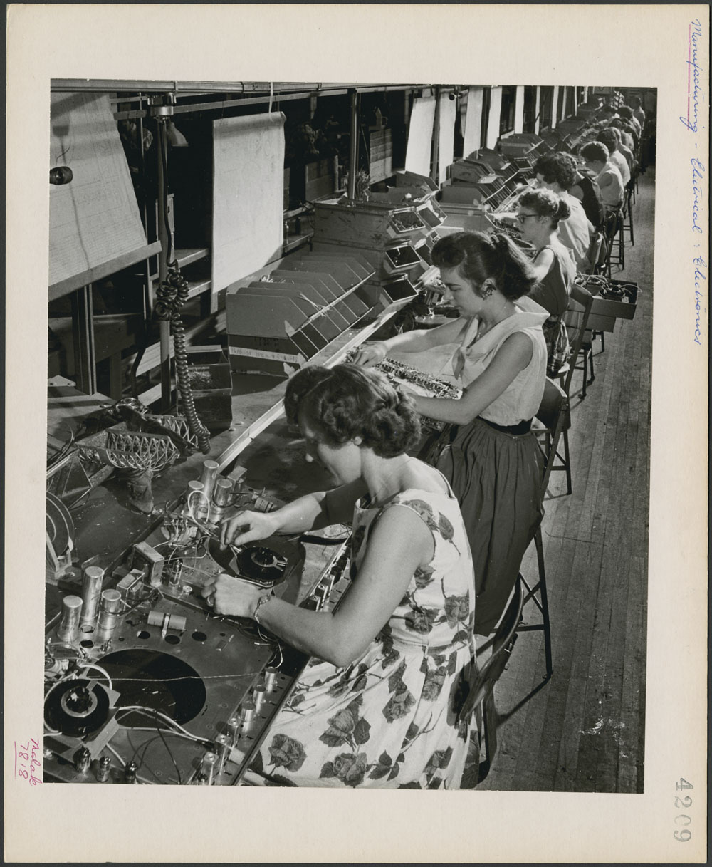 Assembly line of seated women, working on electronics.