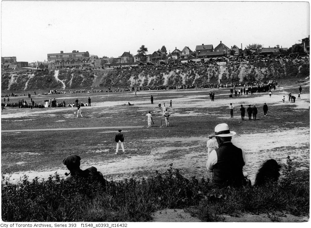 Games of cricket and baseball occurring concurrently in a park. Spectators ring the field.