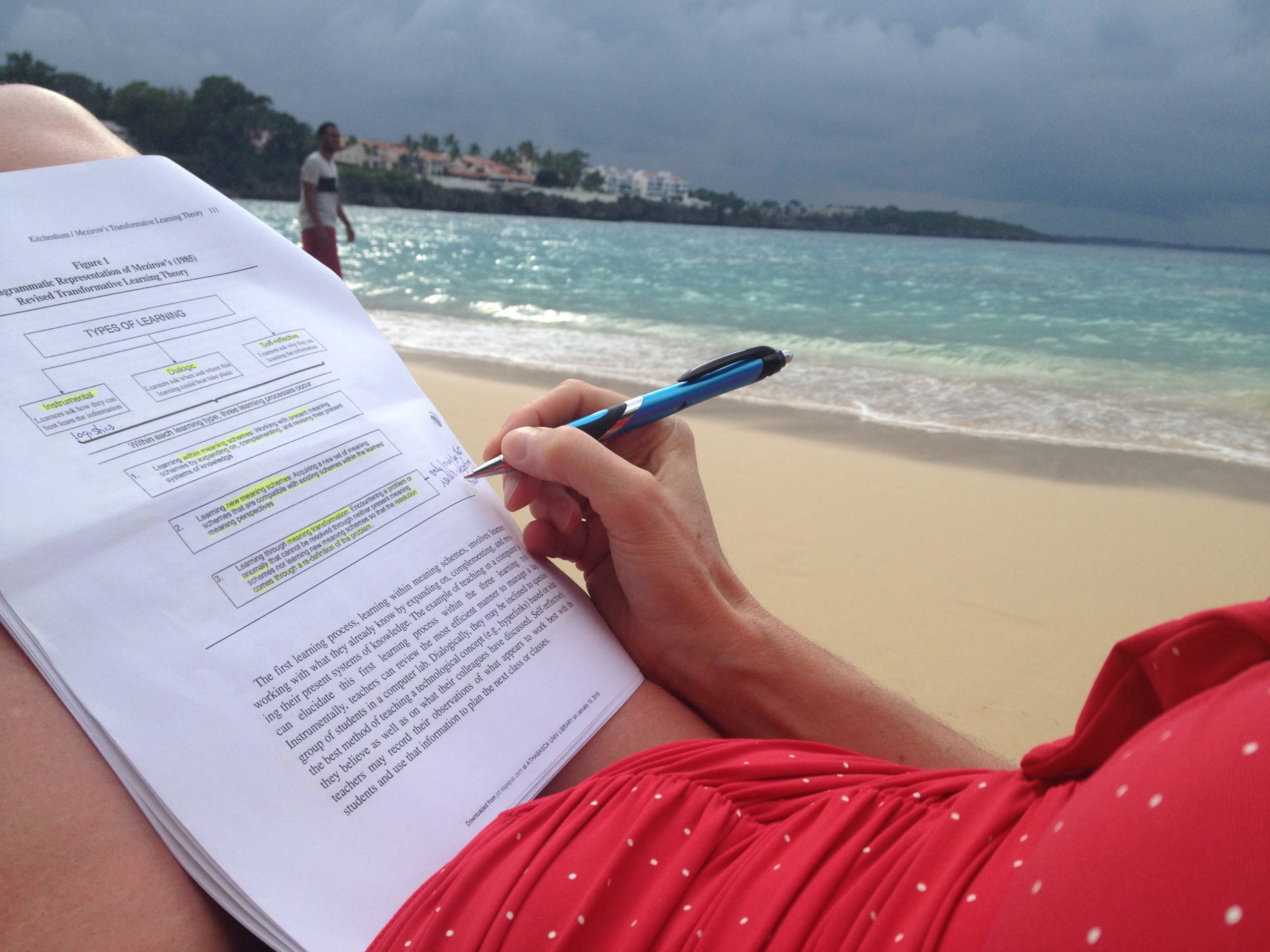 A person making notes on a page balanced on their knees. There is a sandy beach and ocean in the background
