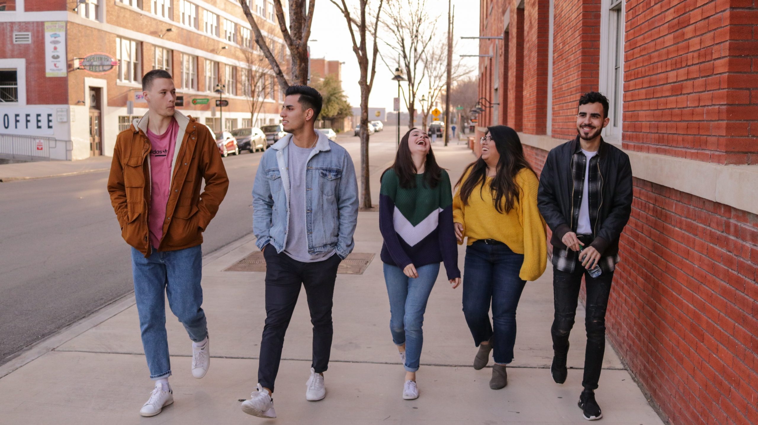 Five college/university-aged students walking down a sidewalk together talking and laughing.