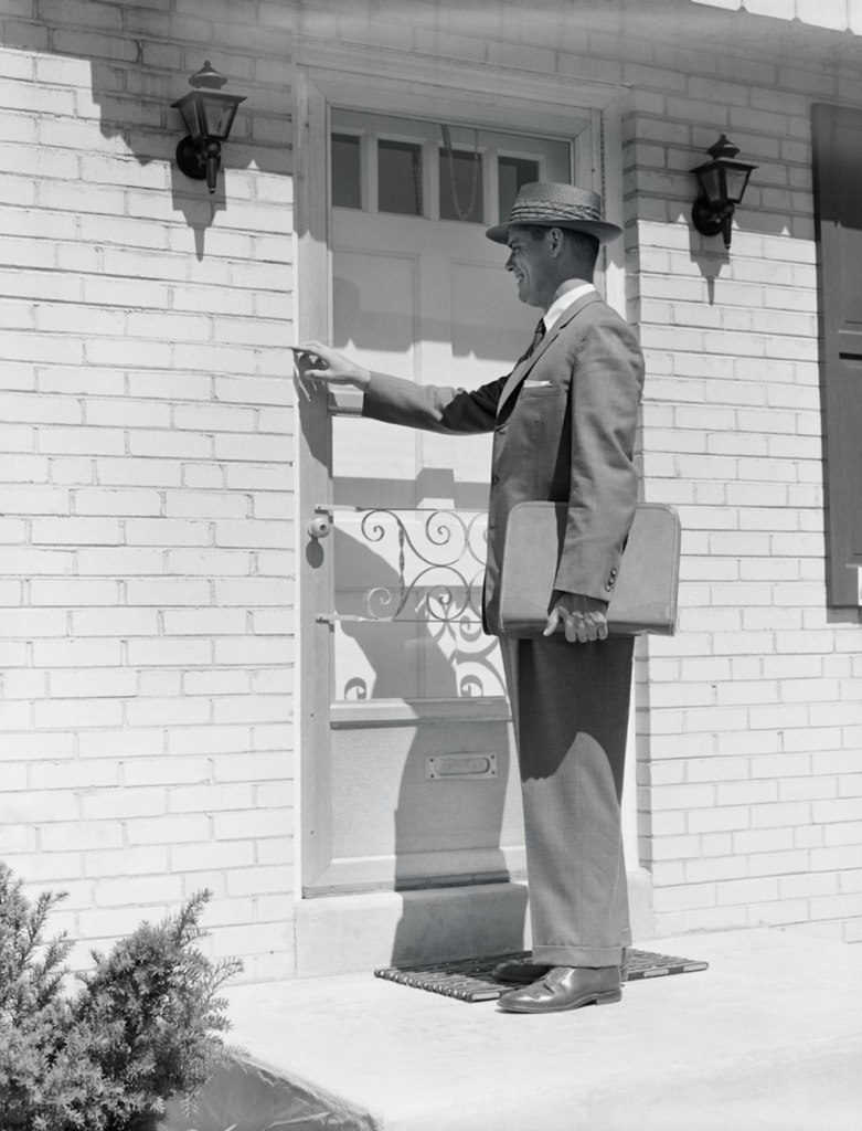 Black and white photo of a man in a suit and hat carrying a briefcase standing in front of a residential door preparing to knock.