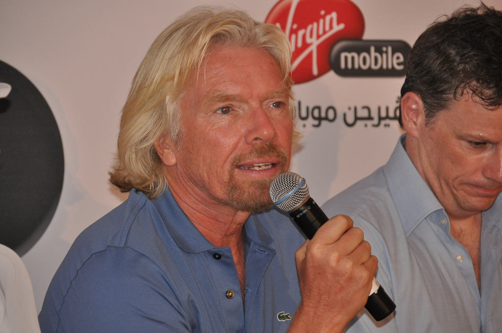 A man speaking into a microphone. A Virgin Mobile logo is behind his head.
