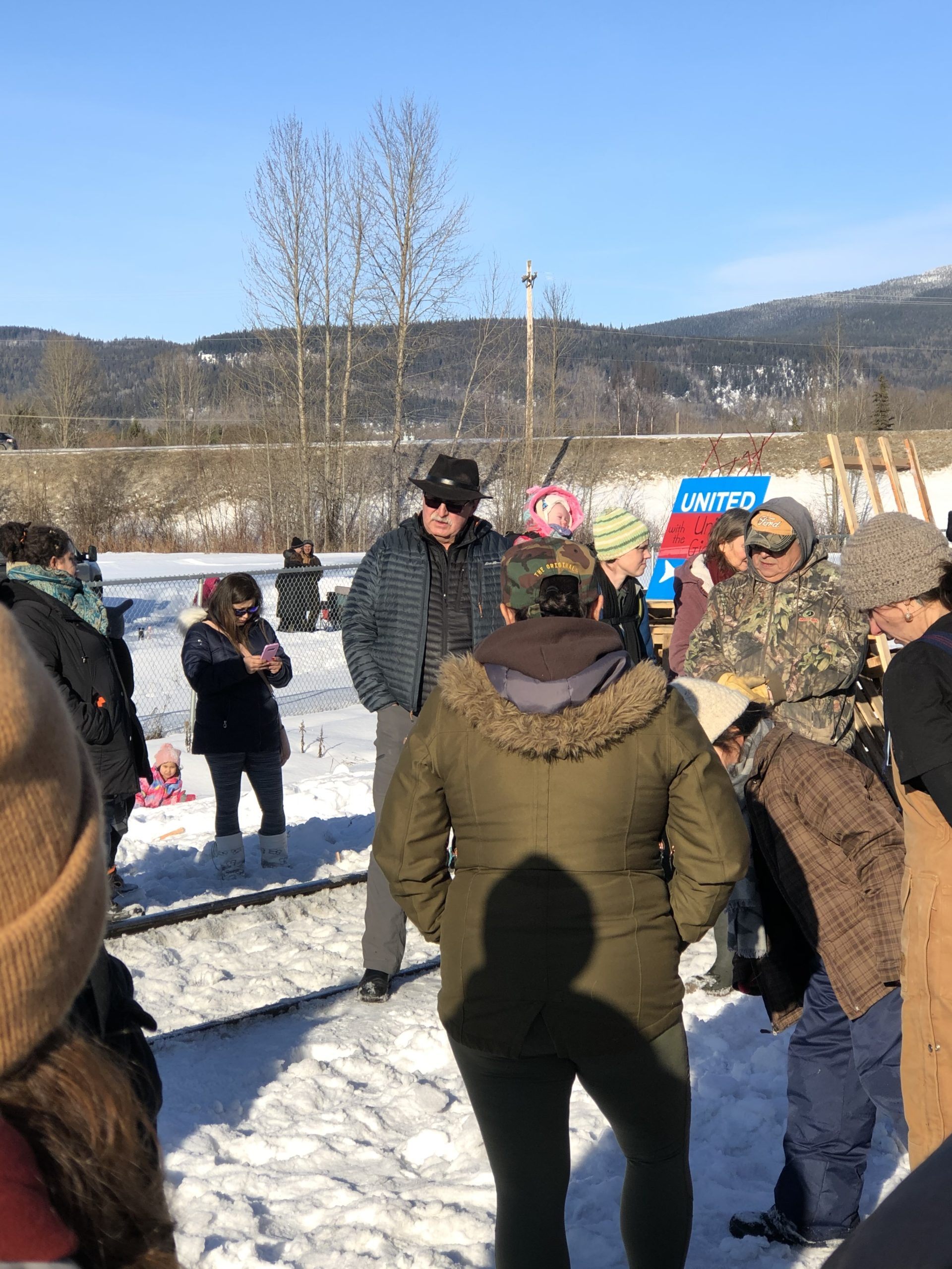 People gathered by a railroad. They stand on snowy ground in winter coats. Some hold protest signs.