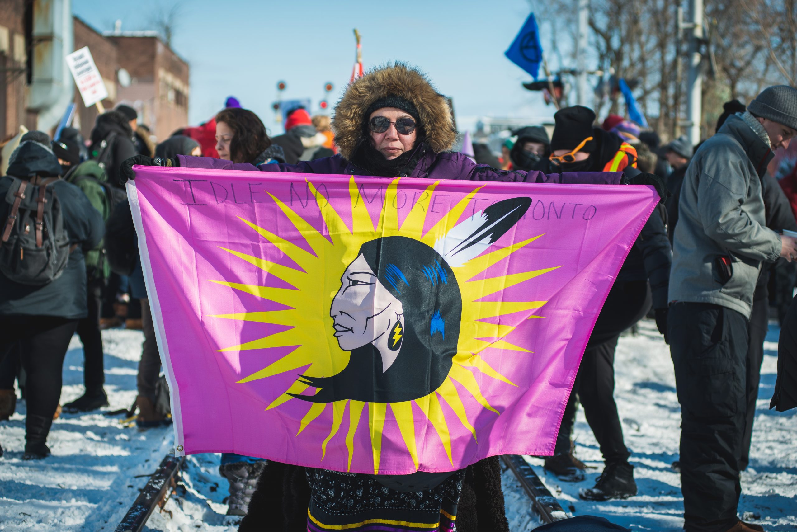 A woman stands in the middle of railroad tracks and holds up a pink flag showing an Indigenous woman wearing a feather in her hair and a lightning bolt earring. The words "Idle No More Toronto" are written in marker across the top of the flag.
