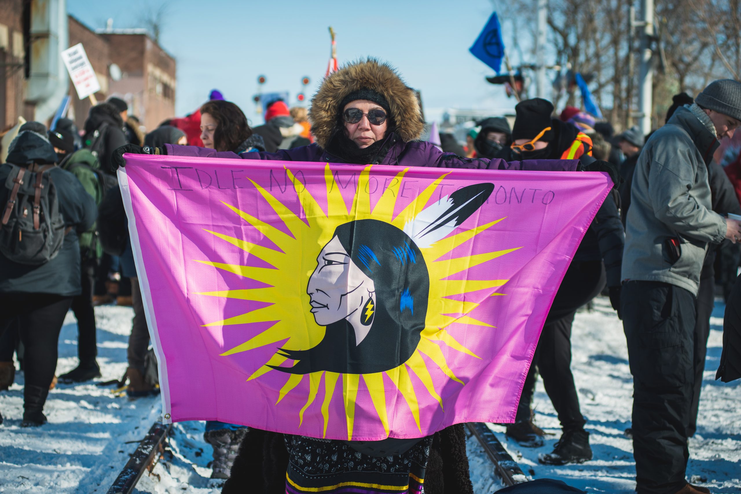 A woman stands in the middle of railroad tracks and holds up a pink flag showing an Indigenous woman wearing a feather in her hair and a lightning bolt earring. The words &quot;Idle No More Toronto&quot; are written in marker across the top of the flag.