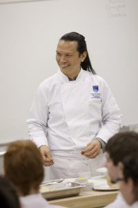 Canadian chef Susur Lee stands at the front of a classroom demonstrating a cooking technique.