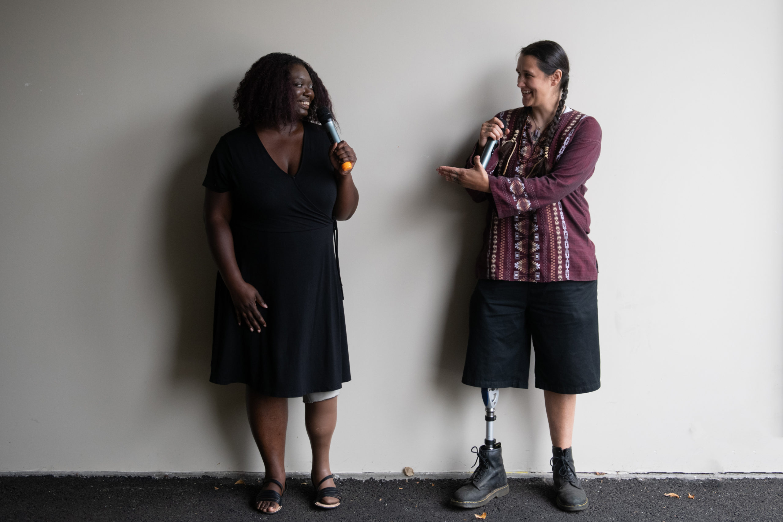 An Indigenous Two-Spirit person and a Black woman face each other and gesture while smiling and speaking into microphones. They are both wearing prosthetic legs.