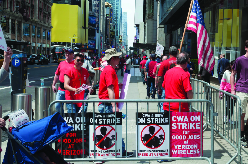 This image shows a group of people dressed in matching red shirts in an area enclosed with signs that say “CWA on strike for good jobs at Verizon Wireless” and “CWA and IBEW on strike. Fighting corporate greed at Verizon.”