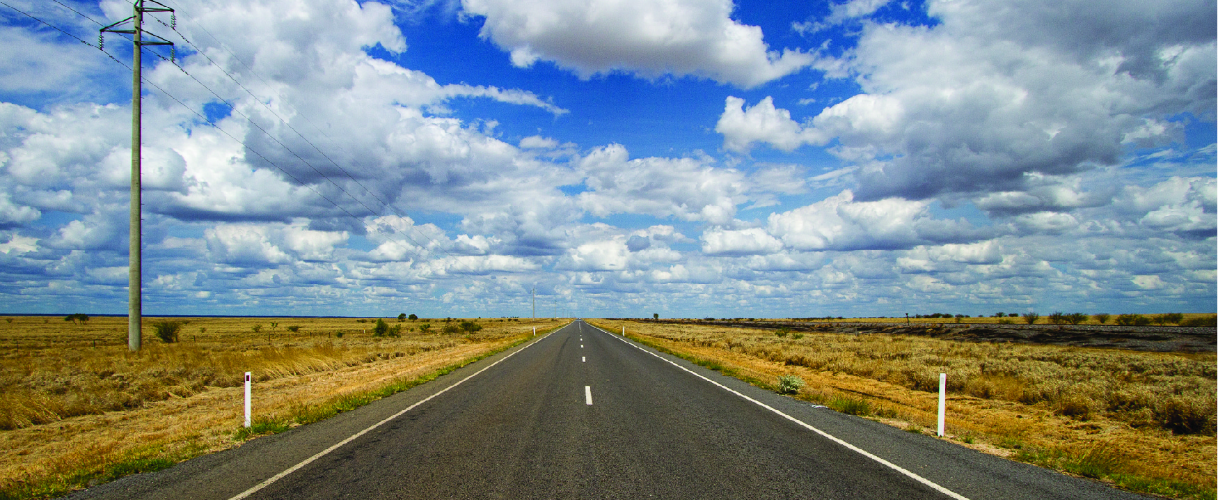This image looks down the middle of an open road with clouds in the sky and open landscape on either side of the road.