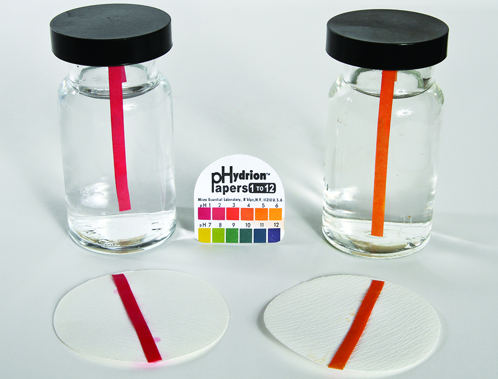 This image shows two bottles containing clear colorless solutions. Each bottle contains a single p H indicator strip. The strip in the bottle on the left is red, and a similar red strip is placed on a filter paper circle in front of the bottle on surface on which the bottles are resting. Similarly, the second bottle on the right contains and orange strip and an orange strip is placed in front of it on a filter paper circle. Between the two bottles is a pack of p Hydrion papers with a p H color scale on its cover.