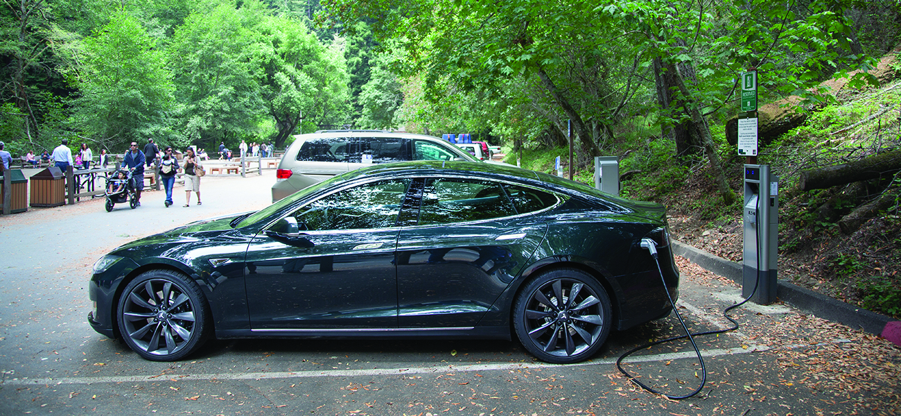 A photograph is shown of a parked car plugged into a charging station in a paved parking area. The parking area is situated in a wooded area. People are walking in the background in the park-like atmosphere.