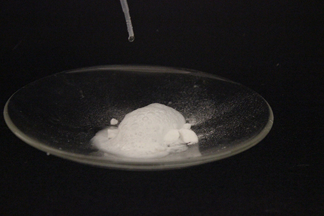A photograph of a watch glass full of a white solid is shown. A plastic pipette drips a colorless liquid into the solid, causing bubbles.