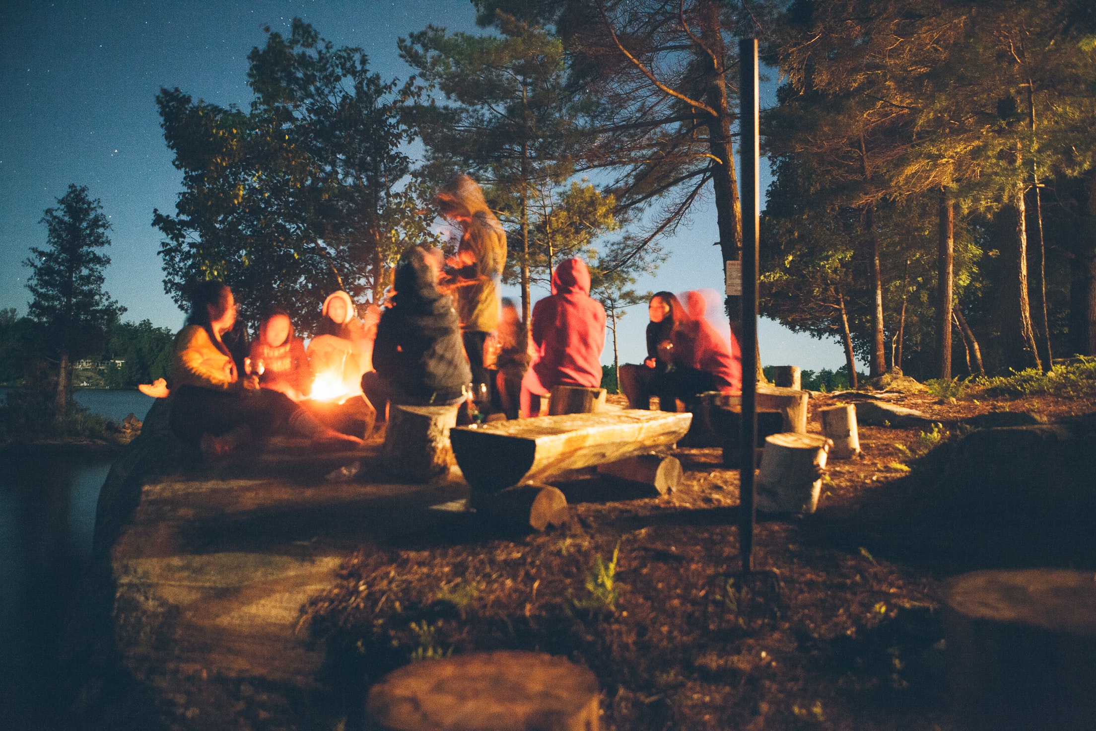A group surrounding a campfire with one person standing and speaking to the group.
