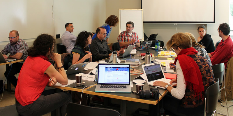 A group of people sitting around a large table talking to each other. The table is filled with books, laptops, sticky notes, and drinks.