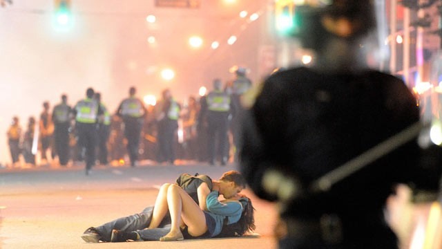 Two people lie in the middle of the road kissing. Riot police are in the foregroud and background.