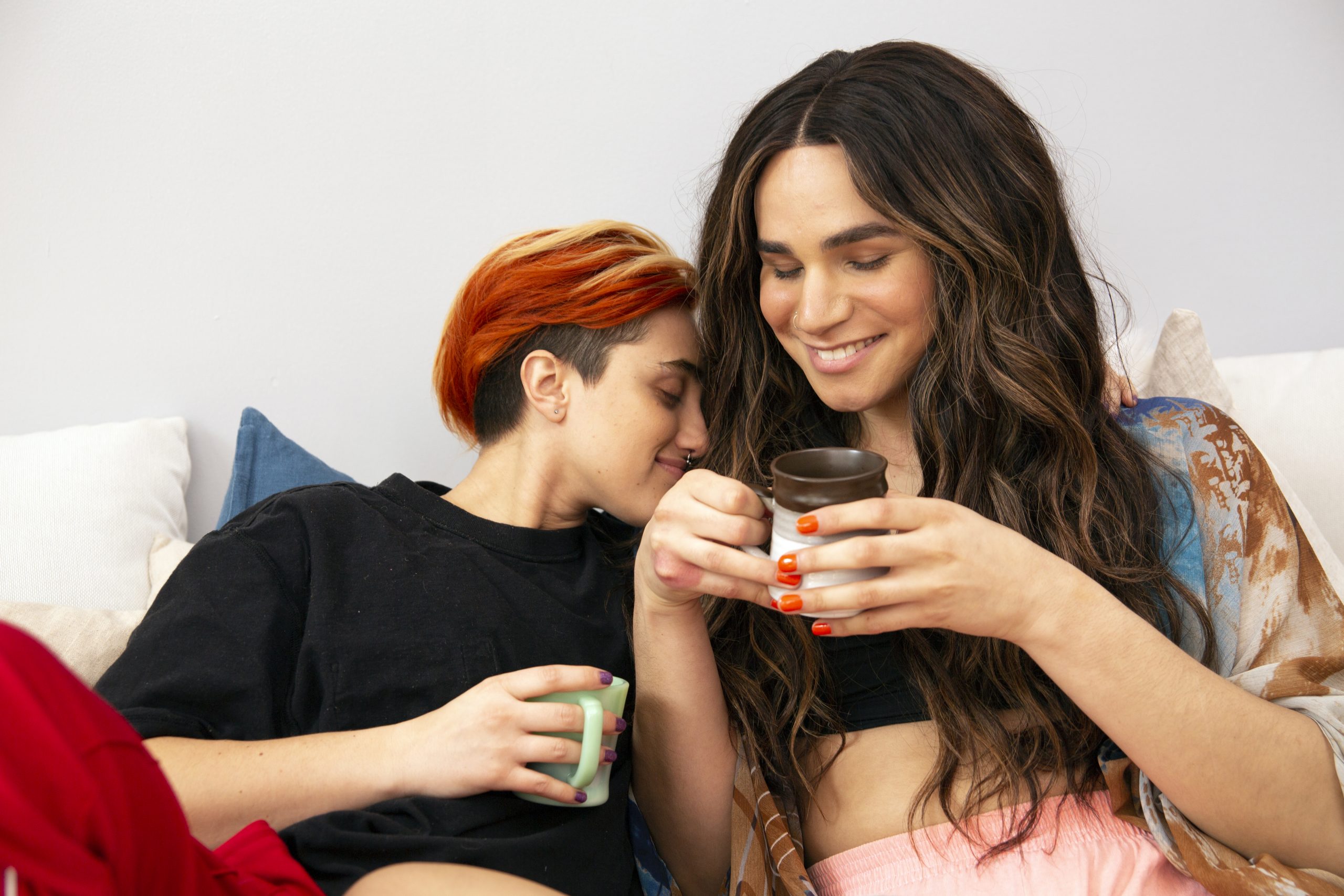 A trans-masculine gender non-conforming person and transfeminine nonbinary person drinking coffee in bed
