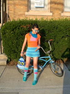 A young woman leans against an old-style blue bike. She wears bright clothes, large glasses, knee high socks and an owl backpack.