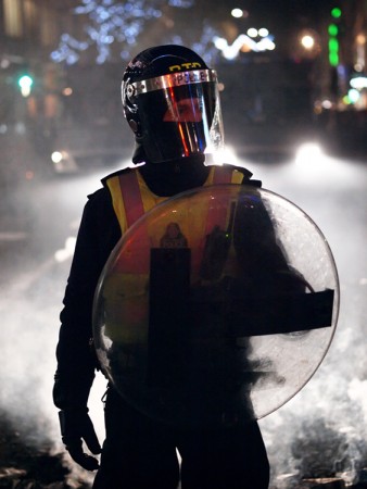 A police officer wearing riot gear including a helmet, a bright vest, and a shield.