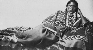 Black and white 19th century, ethnographic style photograph of an Indigenous woman laying on a bed facing the camera.