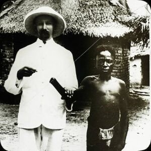 A missionary wearing a light safari suit and a pith helmet, and a Congolese man wearing a loin cloth. The missionary holds up the man's arm at the elbow, and points to the missing hand.