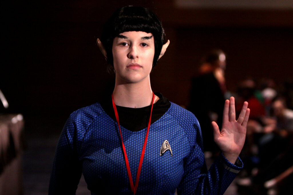 Female Star Trek fan dressed as a vulcan in a blue Federation officer uniform and making the "live long and prosper" gesture with her left hand