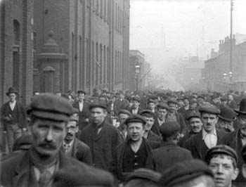 A street full of 19th century industrial workers walking shoulder to shoulder facing the camera as they leave a work shift.