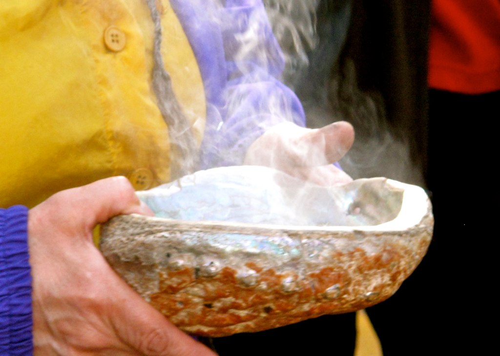 Hands holding an abalone shell shaped like a bowl with smoke rising out of it.