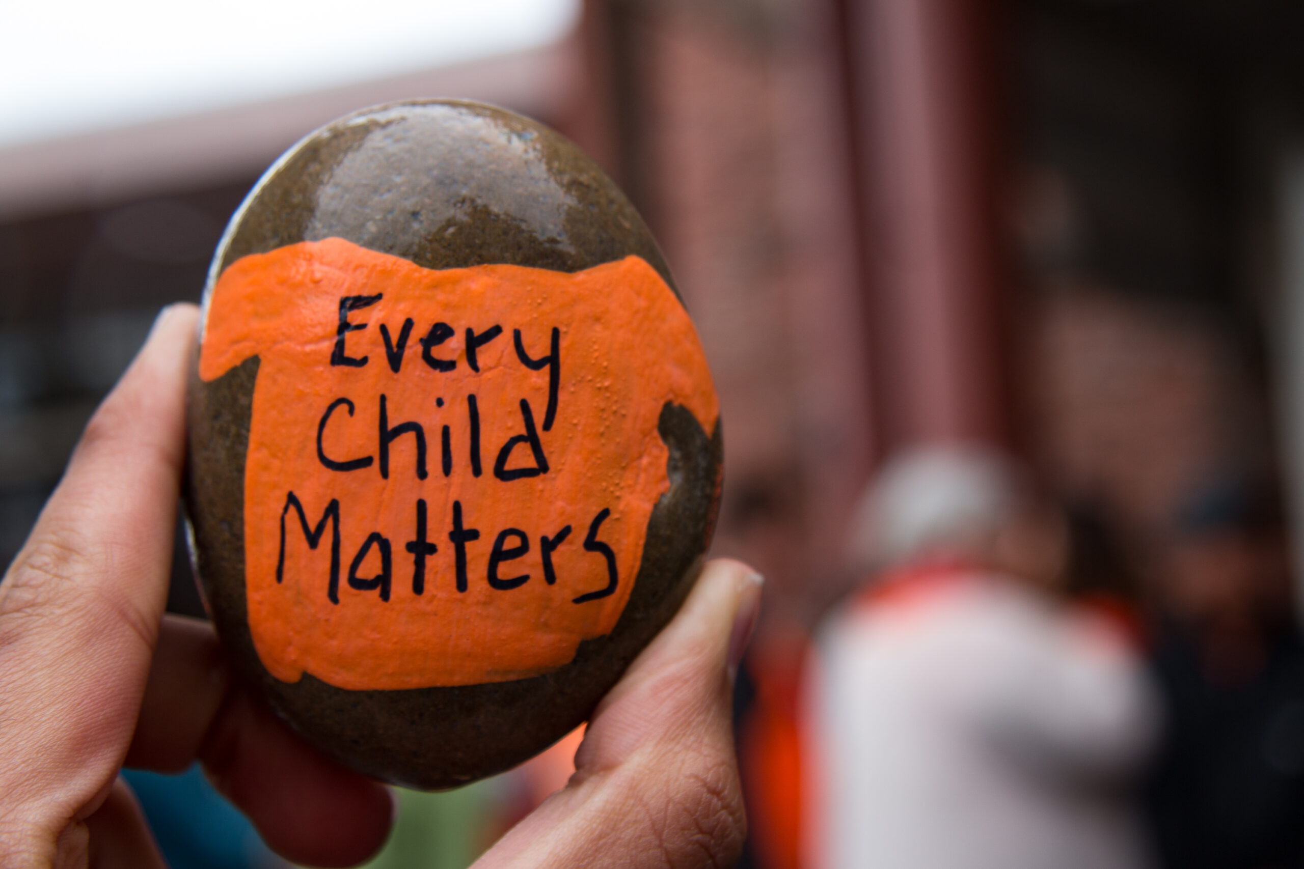 A hand holds up a rock painted with an orange shirt that says, "Every Child Matters."