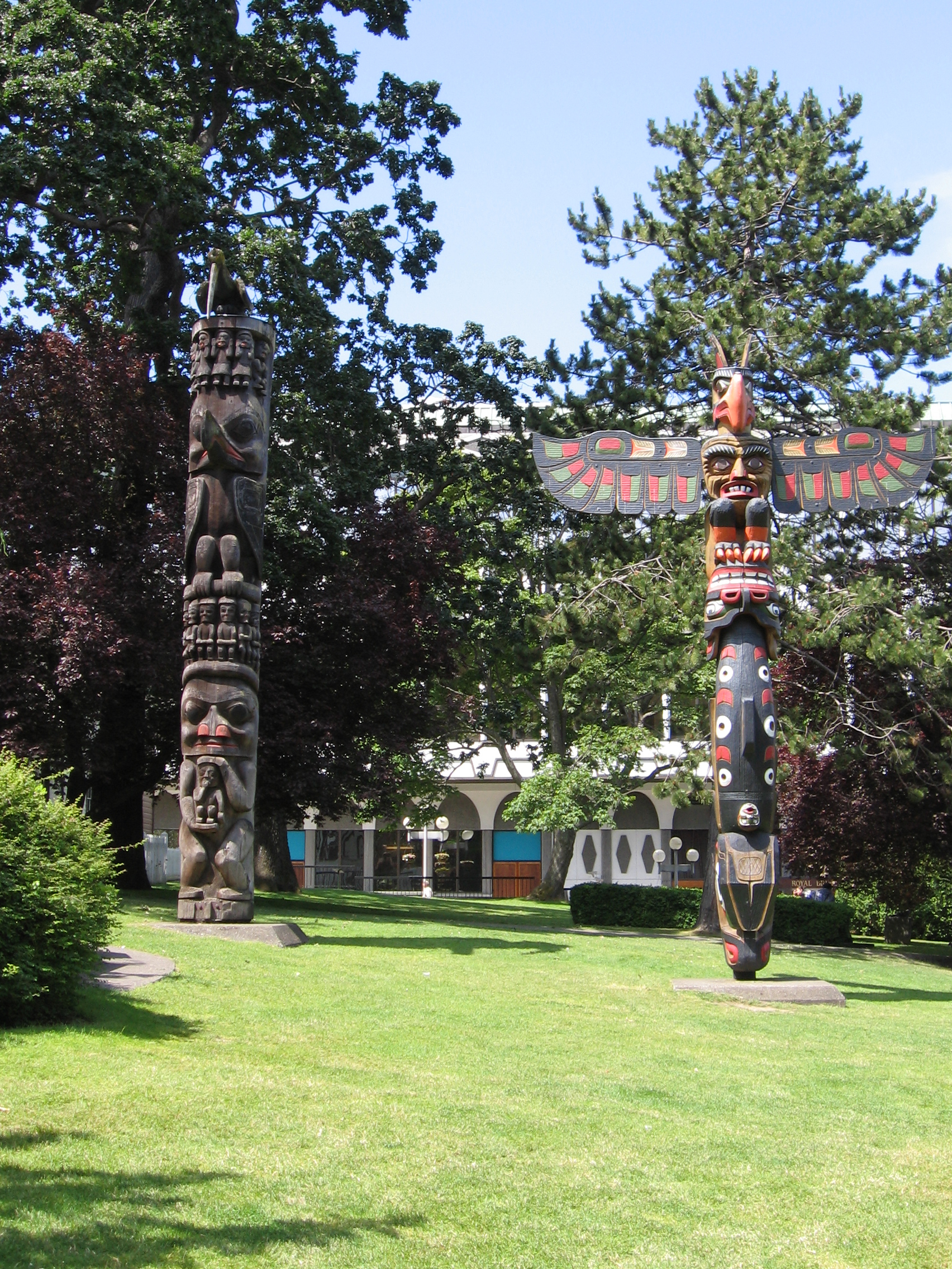 Two totem poles face the camera. In the background, some trees obscure a building.