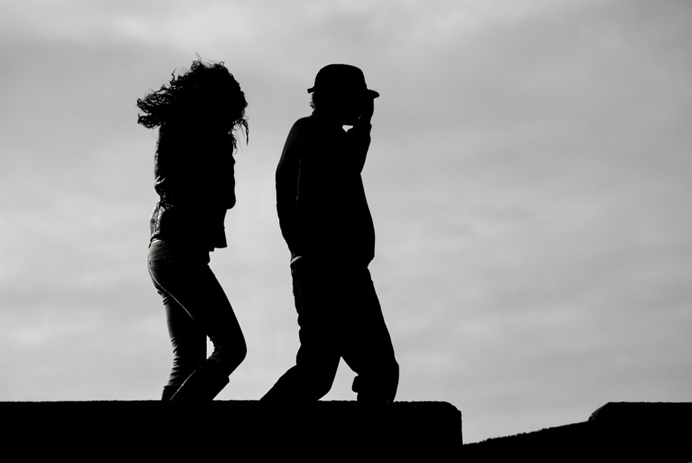 A man and woman are shown walking in the wind, man in front of woman.