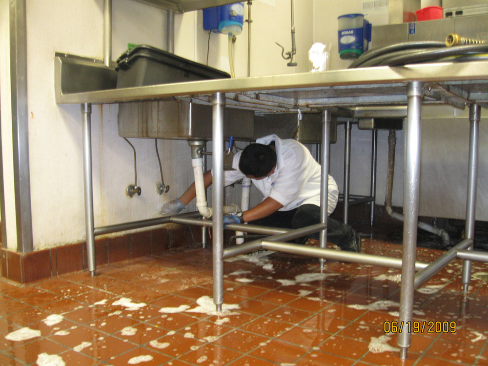 A man is shown scrubbing floors and walls beneath a group of sinks in a restaurant kitchen.