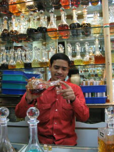 A man stands in front of a tall shelf of glass perfume bottles. He pours perfume from a large bottle into palm-sized bottles.