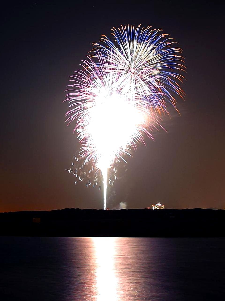 Fireworks explode in the night sky. A reflection of the fireworks is seen on water below.