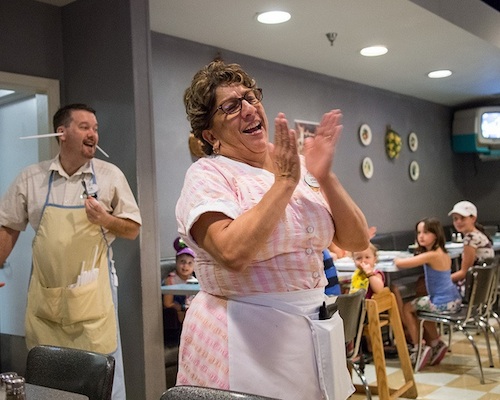 A woman in an apron sings and claps, a man behind her smiles with straws sticking out of his ears.