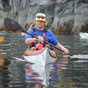 A man wearing a baseball cap with sunglasses balanced on the brim paddles a kayak.