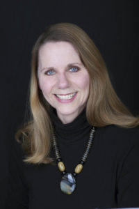 A woman in a black turtleneck and a shiny stone necklace smiles for a portrait.