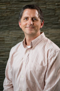 A middle-aged man in a collared shirt smiles in front of a stone wall.