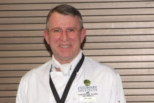 A middle-aged man in a chef’s jacket smiles in front of a wall.