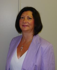 A woman with short hair wearing a lilac blazer poses in front of a white wall.