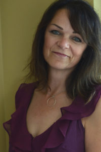 A middle-aged woman poses wearing makeup and a ruffled purple dress.