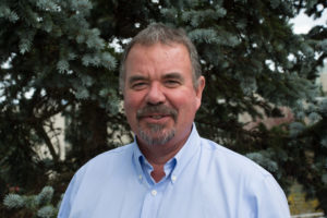 A smiling middle-aged man in a collared shirt poses beside a coniferous tree.