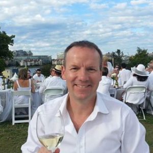 A man in a white shirt holds a glass of wine. He dines outside with other people dressed in white.