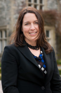 A woman in a blazer smiles and poses. In the background is a stone building.