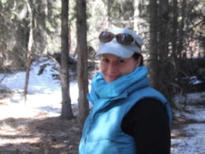 A woman wearing a puffy vest stands in a sunny forest with scattered snow on the ground.