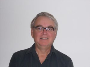 A middle-aged man in glasses and a collared shirt poses in front of a blank wall.